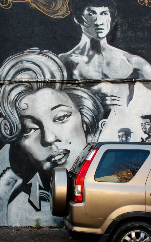Street art painting in grey tones, a topless Bruce Lee  is standing behind the head and shoulders of Marilyn Munro. 