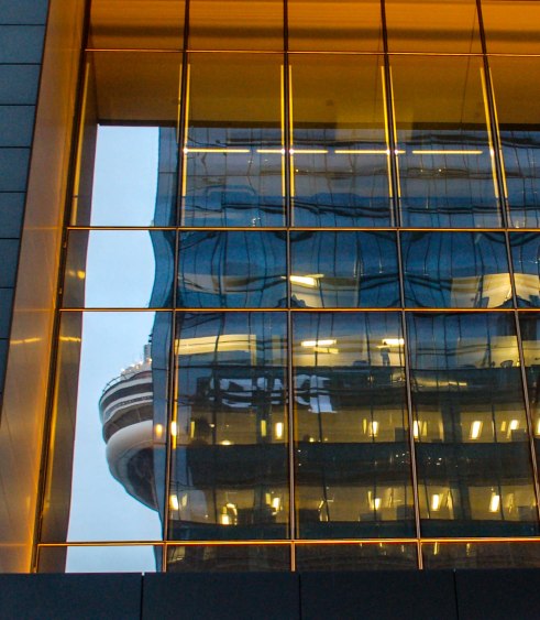 Early evening, the CN tower is refleceted in an office building.  The lights are on and you can see people working in the building. 