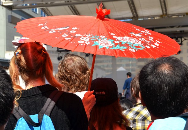 A woman holding a red paper umbrella standing beside a woman wearing a baseball hat that says Toronto on it. 