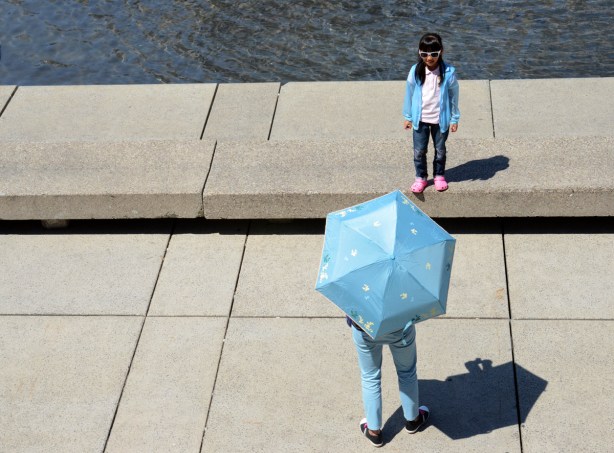 A girls wearing a light blue jacket is standing beside the pool at Nathan Phillips Square while her mother takes her picture. The mother is wearing light blue pants and is holding a light blue umbrella. 