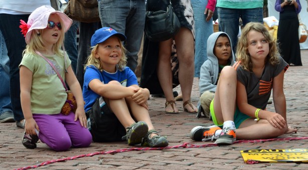 four children sitting on the ground and watching a magic show. Three are smiling, one is looking skeptical.