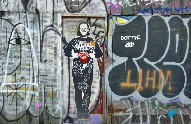 Black and white graffiti on a wall and door.  There is life sized picture of a man on the door.  He is holding a red heart in his hands.  