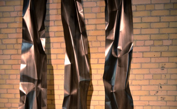 A sculpture that is composed of three tall vertical pieces of shiny metal. A yellowish brick wall is in the background.