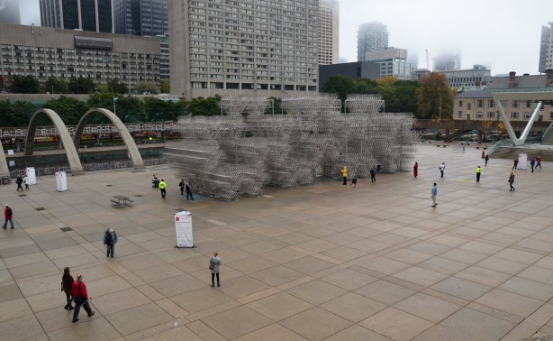 Nathan Phillips Square on a grey and foggy morning.  There are a few people in the square who are looking at the large art installation that involves a structure made from more than 3000 bicycles
