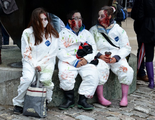 three zombies dressed in white coveralls, sitting by The Archer sculpture in Nathan Phillips Square