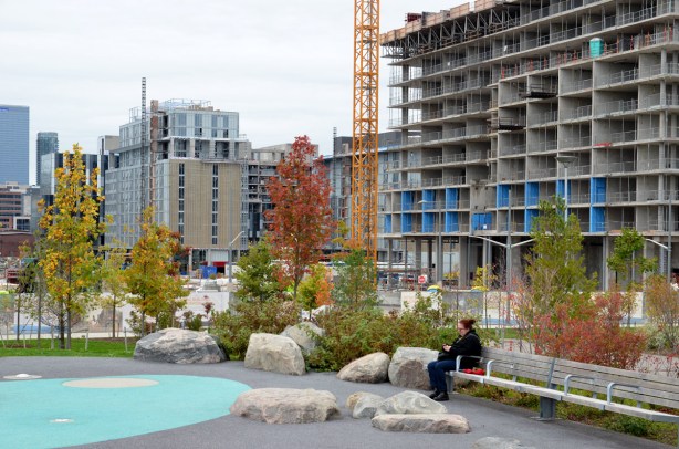 a woman is sitting on a bench, looking at her phone.  Behind her there are a number of buildings under construction