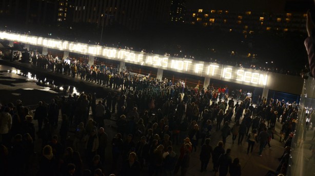 Crowds, at night. Light is provided by fluorescent lights that spell out a short poem.