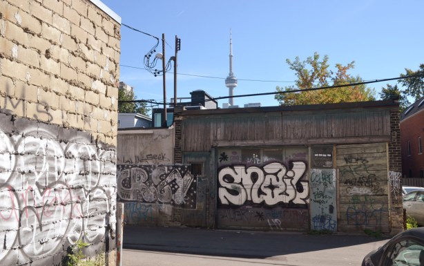 buildings in an alley that have graffiti on them.  the CN Tower is in the background