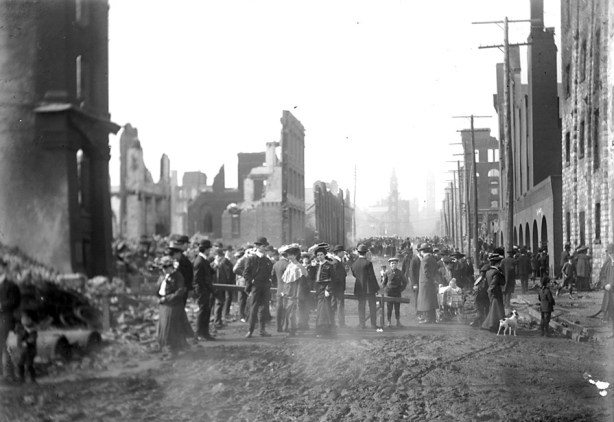 Historical photo of Bay street after the 1904 fire in Toronto.  There are people on the street.  The street is all mud.  There are many burned out buildings on both sides of the streets.  Brick facing of two to four storey buildings is all that remains.