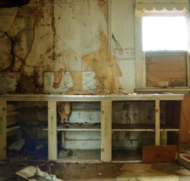 one wall, interior, of a kitchen in an abandoned building.  The window is half boarded up.  The cupboard doors are missing.  The wallpaper is almost all peeled off.  It is filthy.