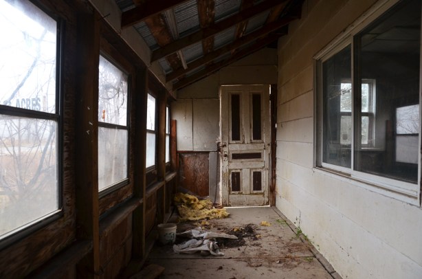 The interior of the front porch room of an abandoned house.  There is lots of debris on the floor, no curtains on the windows. 