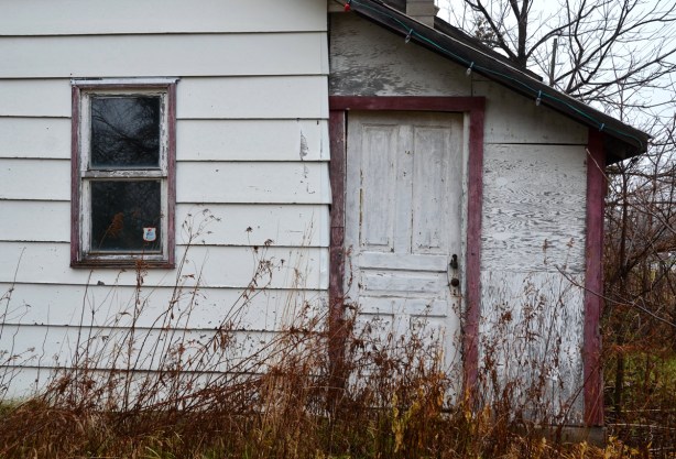 The side of an old white frame house.  The red trim is looking very faded.  The grass in the yard is very long. 
