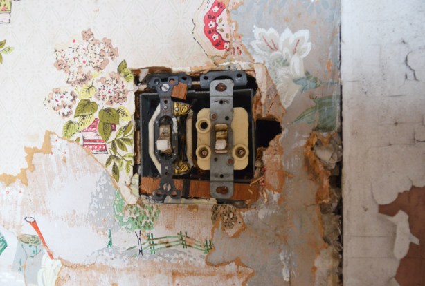 A double light switch that has lost its cover plate, on a wall.   The wall paper around it has been torn in such a way that 3 layers of different floral paper can be seen. 