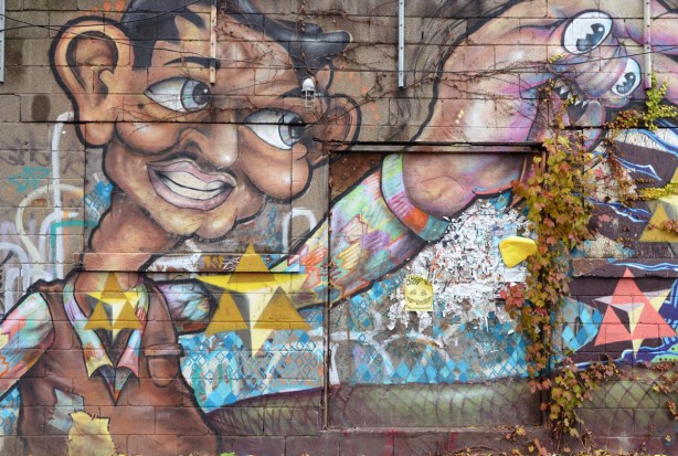 mural showing a man's upper body and arm. The head and hand are oversized. The hand has been made to look like a head, with eyes on two of the fingers and a mouth with teeth on the palm of the hand.