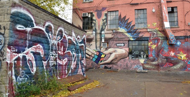 At the intersection of two alleys. Straight ahead is a building covered with a large mural with large hands and a Rubik's cube amongst other things. On the right is a one storey building with a poser tag in blues and maroons.