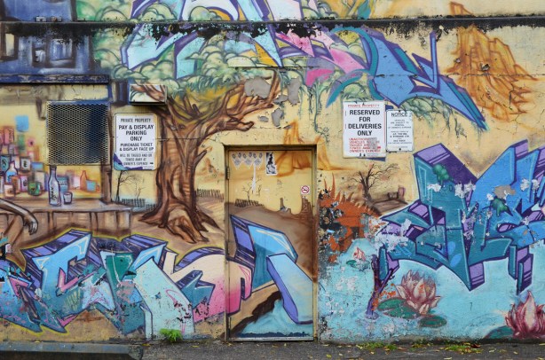 part of a mural on a wall. There is a door in the middle. Part of the mural shows a large tree with a brown trunk and multicoloured leaves.