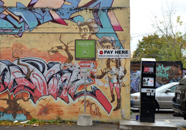 part of a mural on a wall. It is also a parking space for zip cars. The mural has a pale yellowish background. It has blue an dpink tags as well as a picture of a man.