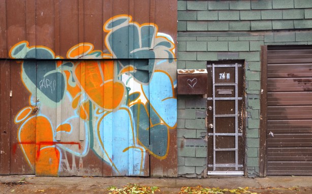 brown garage door, brown front door and rusty brown mail box.  On the garage door is a piece of street art with orange and blue blobs.  The front door is decorated with a grid made of metal strips.