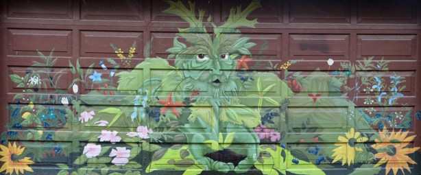 Mural on a garage door showing a man like figure made of leaves and other greenery.  He is surrounded by, of made up of,  flowers and plants