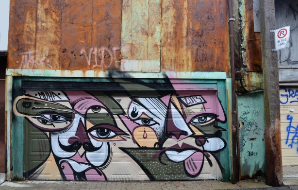 large piece of street art on a garage door, in purples and greys.  Above the garage door is a rusted metal wall. 