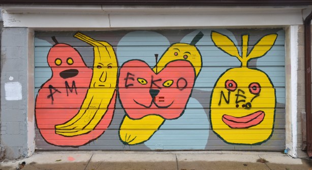 Street art on a garage door showing large stylized fruit with faces on them.  A yellow banana, an orangish pink pear and apple and two other pieces of yellow fruit.