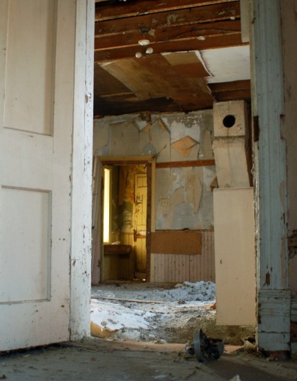 Looking through an open doorway into a room that has a layer of debris on the floor.  The ceiling has fallen (or been removed) and just the wood can be seen. 