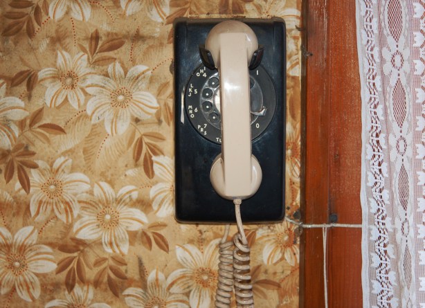 an old rectangular rotary phone hangs on a wall that is covered with brown, beige and white floral wallpaper.  The phone is black but has a beige handset.