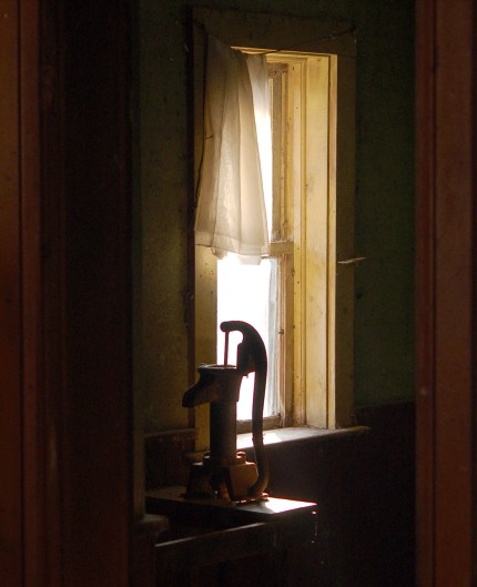 On a counter there is an old hand pump (or water).  It is backlit by sunlight coming through a window.  There is part of an old sheer curtain covering the top part of the window