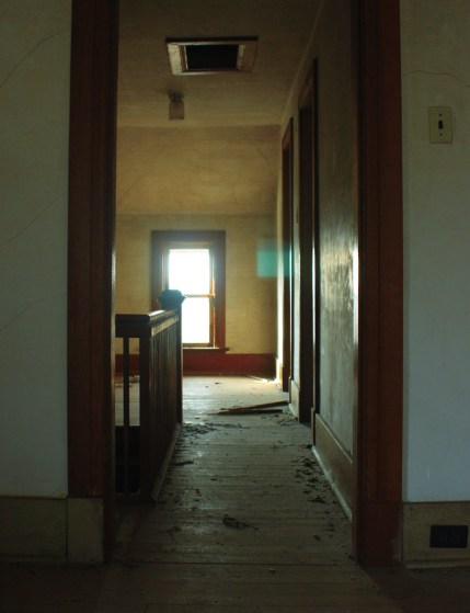 looking down a hallway towards a window.  There is a bannister on the left and a couple of doorways on the right.  The floor is dirty.
