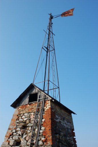 The windmill.  Stone and brick building, pump house, below with a metal frame teepee shaped structure on top.