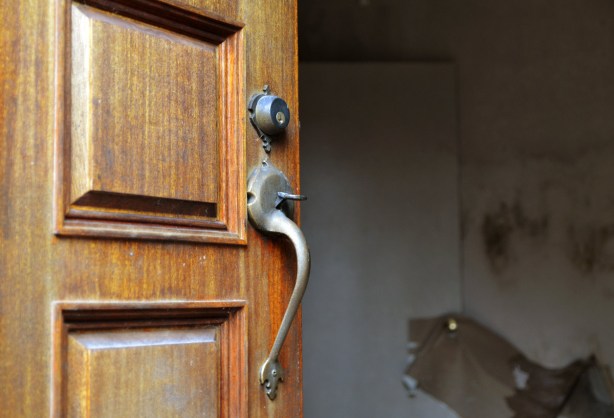 close up of a wood door showing the middle section of the door with a metal lock and handle.  The interior of the house is barely visible but it looks grey inside. 