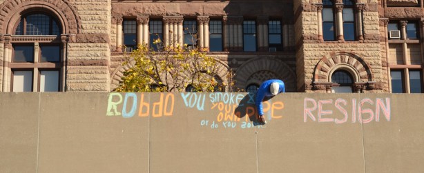 A man is writing in orange and blue chalk.  Old City Hall is in the background. 