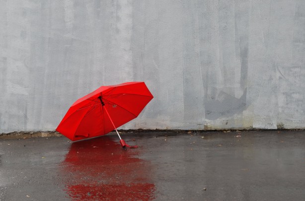A red umbrella is open and sitting on the wet pavement beside a grey painted wall.