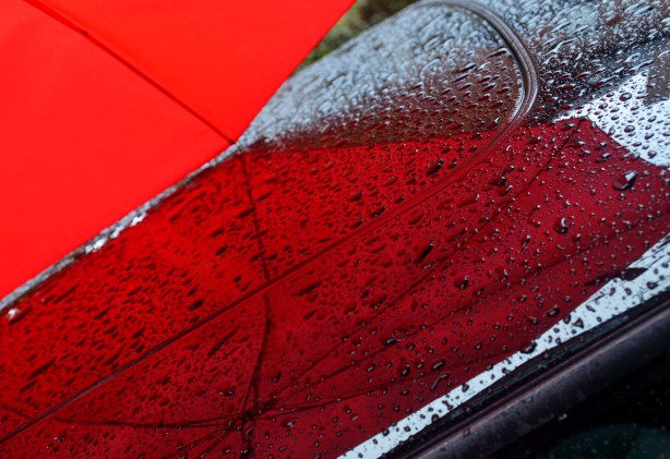 A close up photo of the red umbrella's reflection on the roof of a black car. There are lots of rain drops on the roof as well.
