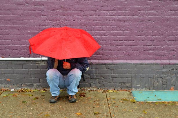 A man is crouching beside a brick wall that has been painted purple and grey. He is holding a red umbrella over his head.