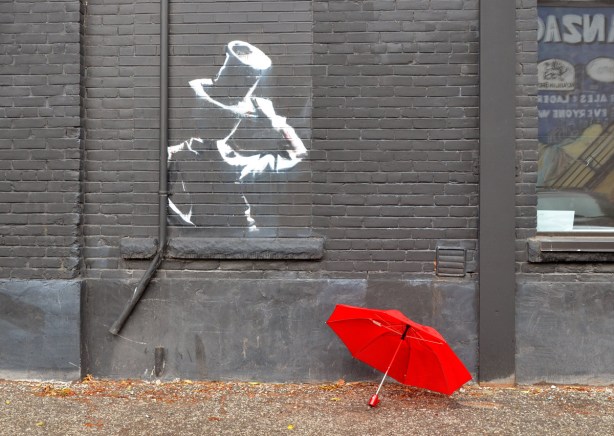 A white line drawing of a man with a top hat (upper body only) on a dark grey wall. On the ground beside the wall is a red umbrella