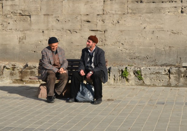 two older Turkish men sitting on a bench talking to each other.  They are dressed for cold weather