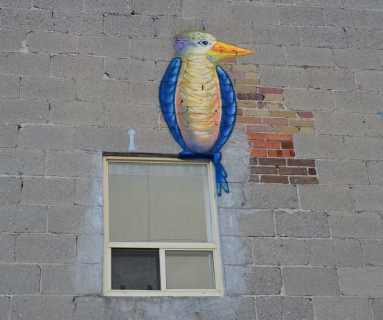 A graffiti painting of a blue and white bird on a grey concrete block wall.   It is painted to look like it's perched on the top corner of a window. 