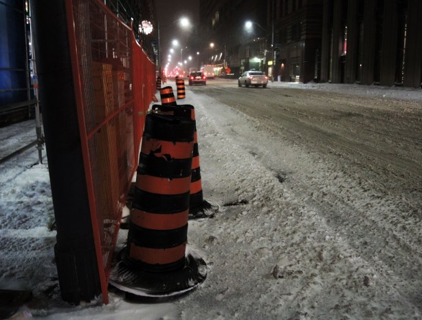 Black & orange construction cones along side a plastic orange fence around a construction site at the side of the road. Traffic stopped at a stoplight in the distance.