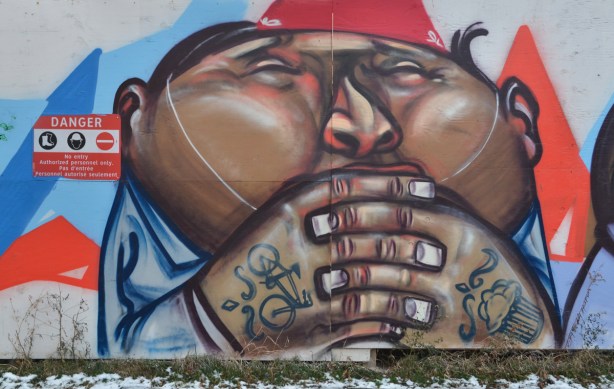 graffiti picture of the face, shoulders and arms of a fat man. The backs of his hands have tattos of a bicycle and a cupcake. 