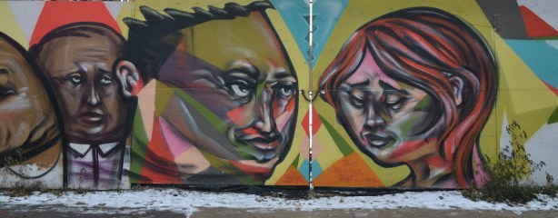 Three faces graffiti. Two men's heads on the left and a woman's head on the right. They are painted on a wood gate that is closed and locked with a chain. 