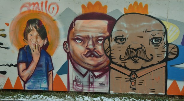 graffiti picture of the upper bodies of 3 people, 2 men and a girl. The girl seems to have an orange halo around her head. The men have orange crowns on their heads. 