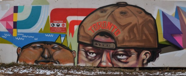 Street art picture of 2 male heads. One is wearing a brown baseball cap backwards. The word Toronto is written on it in orange letters. 