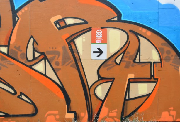 Part of an orange and beige graffiti tag. It surrounds a small sign that points to the entrance to the train station. 