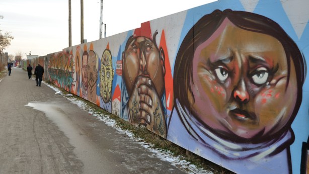 Looking along a temporary wood fence that has been covered with graffiti. The art that you can see in the foreground consists of a number of head. A dirt pathway runs along beside the fence. 