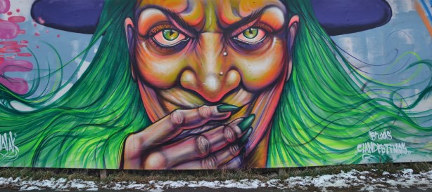 graffiti of a witch's face. She has green eyes and green hair. She is wearing a purple hat. Her boney hand is in front of her mouth so you can see her long green finger nails.