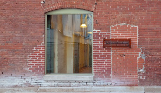A red brick wall with an old door that has been bricked over.  A large new window has been added.  You can see the lights on inside.
