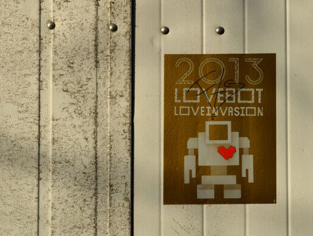 gold coloured poster on a dirty light grey garage door.  The poster says 2013 Lovebot Loveinvasion.  It has a picture of a light grey robot on it, with a red heart. 