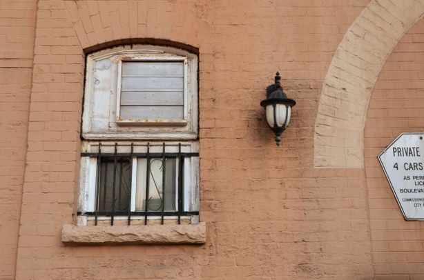 A brick wall that is painted a pinkish colour.  There is an old window with the lower half barred.  A light is mounted on the wall to the right of the window.  The wall is dirty. 