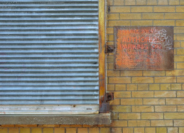 An old metal sign saying No Authorized Parking has rusted badly.  It is on a yellow brick building.  Beside it is a blue coloured metal screen covering a window. 